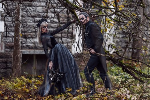 A couple poses in gothic outfits and skull makeup for Halloween in a fall setting.