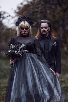 A couple in gothic attire with skull makeup posing in a spooky forest.