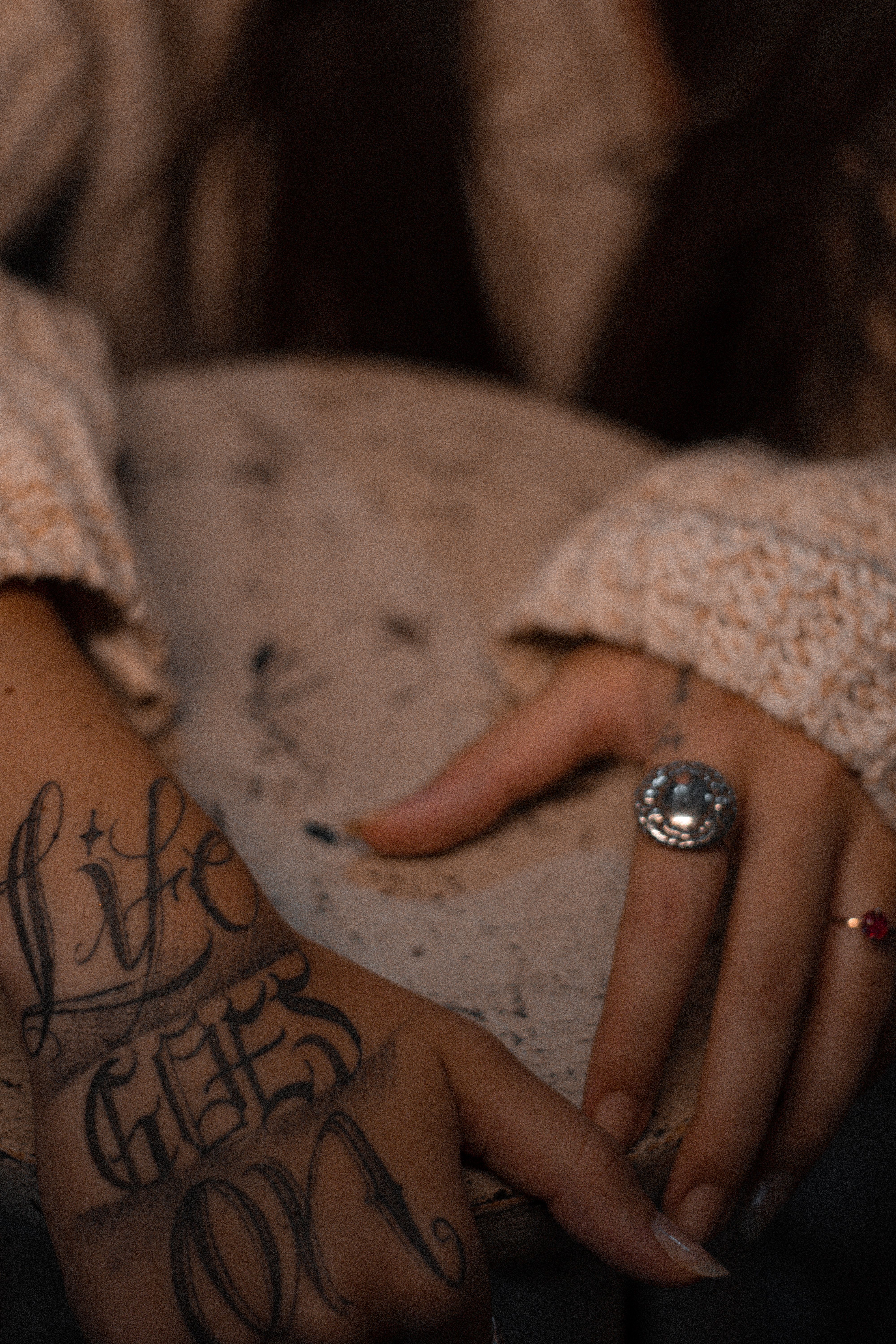 Close-up of a tattooed hand with rings, wearing a knitted sweater, resting on a rustic table.