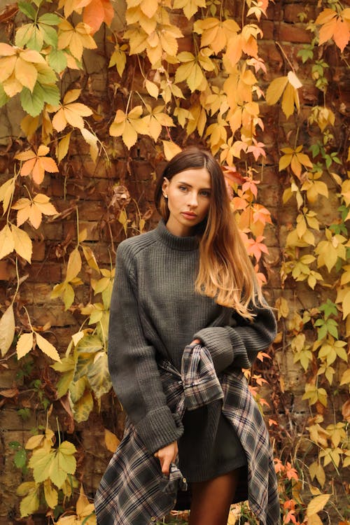 Model Tying a Plaid Shirt Over a Gray Sweater Standing in Front of a Brick Wall Overgrown by Vines