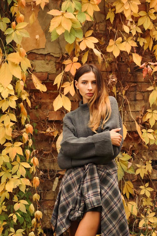Model in a Gray Sweater and a Plaid Shirt Tied Around Her Waist in Front of a Brick Wall Overgrown by Vines