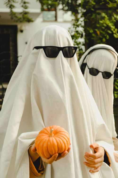 Free Two people in ghost costumes with sunglasses holding a pumpkin, embracing Halloween fun. Stock Photo