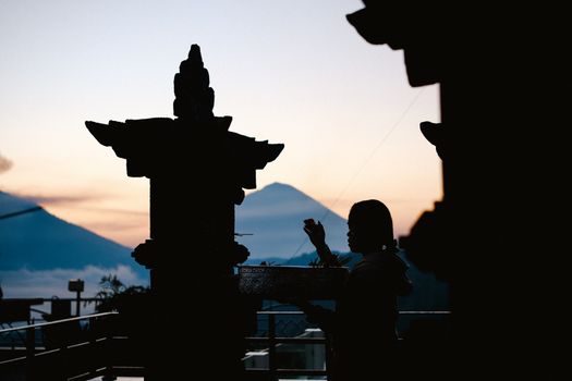 A silhouette of a woman making offerings at a temple with mountains in the background during sunset.