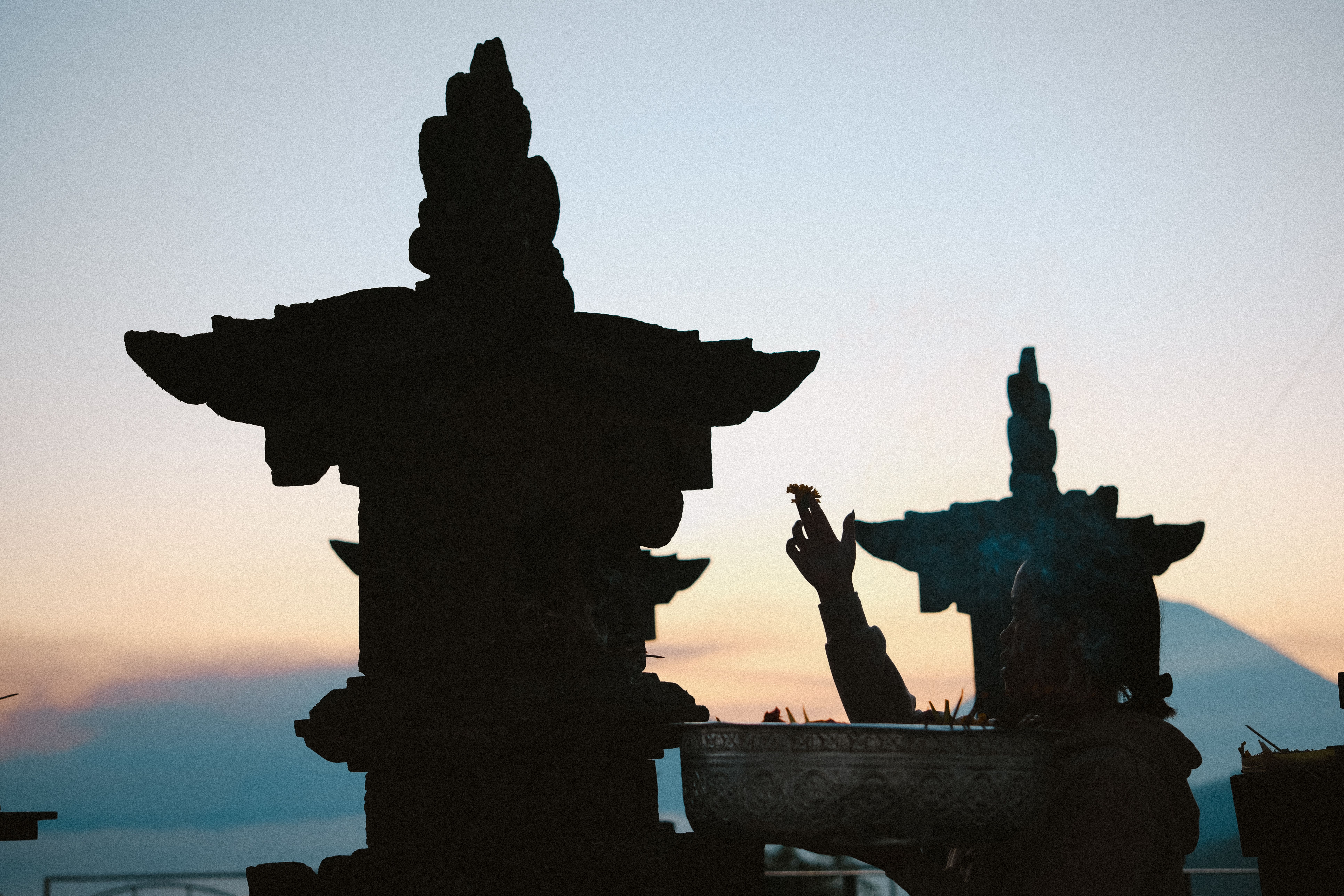 A person performs a ritual at sunset beside temple structures, silhouetted against the sky.