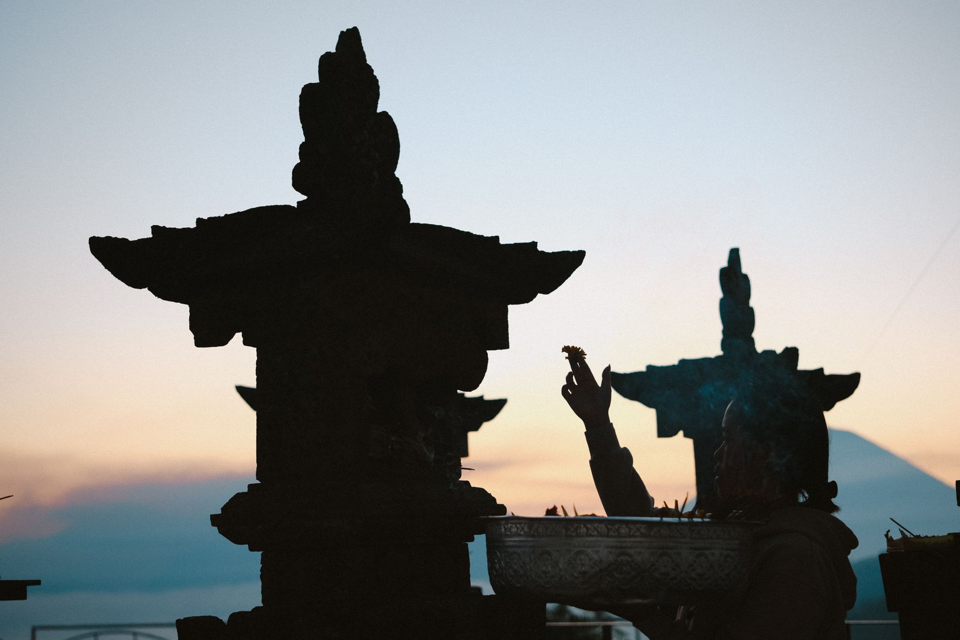 A person performs a ritual at sunset beside temple structures, silhouetted against the sky.