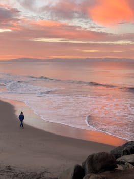 Person walking on Santa Cruz beach at sunrise with vibrant sky and gentle waves.