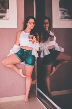Stylish woman in casual summer attire poses by a reflective glass wall holding a cup.