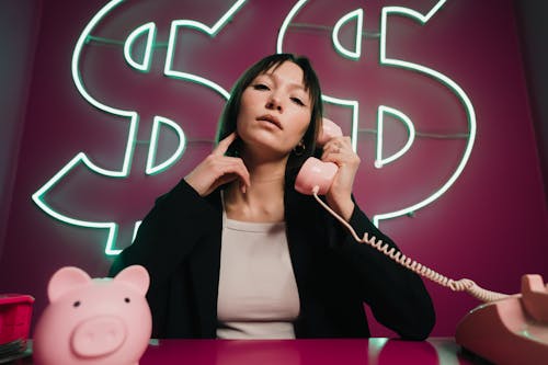 Woman Talking on Telephone Sitting Under Neon Dollar Signs with a Piggy Bank on the Desk