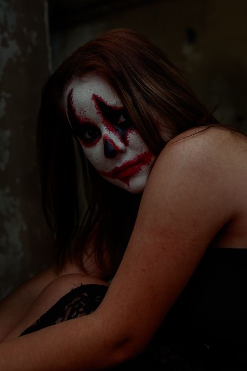 Free A haunting portrait of a woman with horror clown makeup sitting in a dark room. Stock Photo