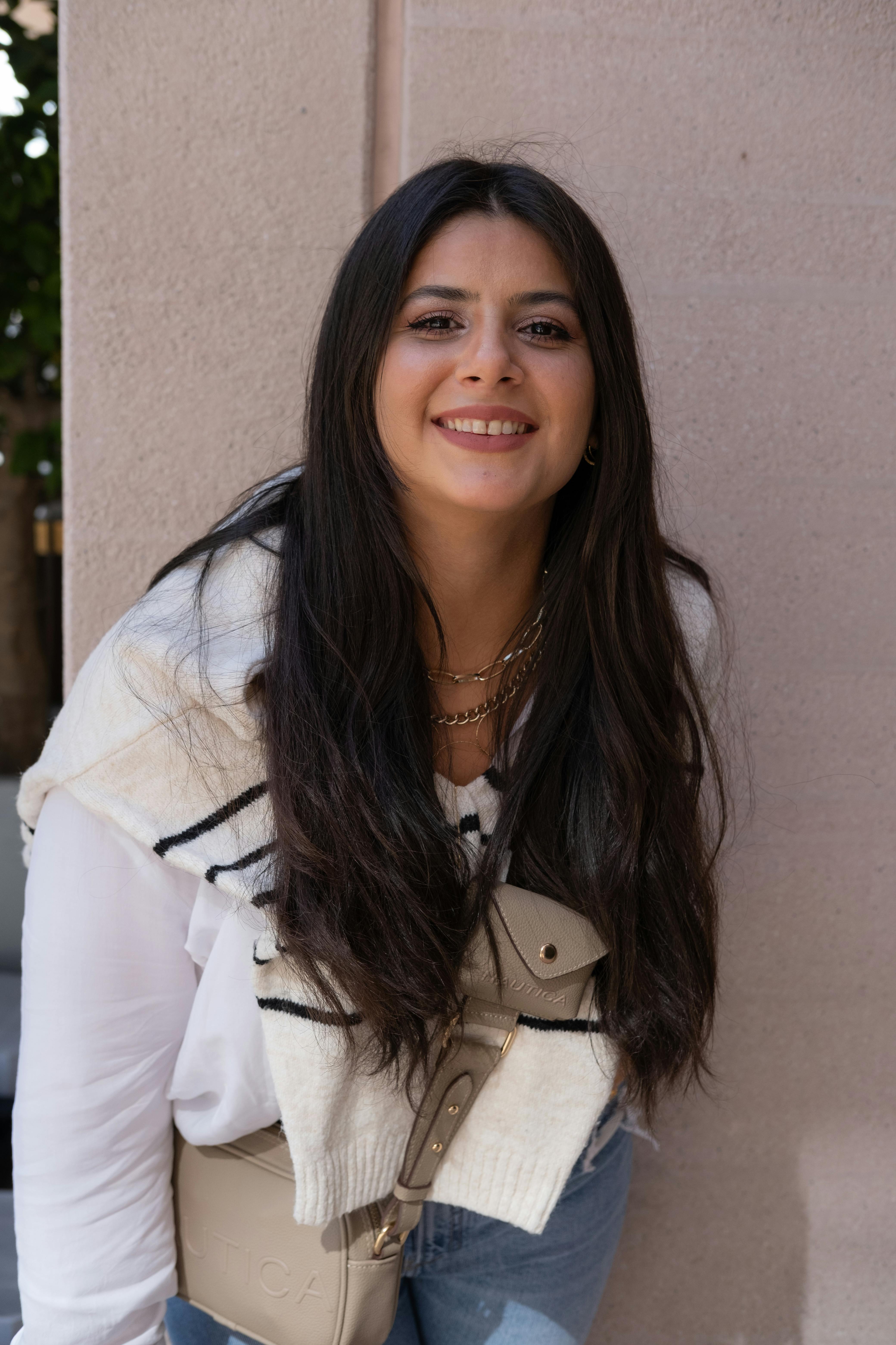 Chic young woman with long brunette hair smiling in stylish outfit outdoors.