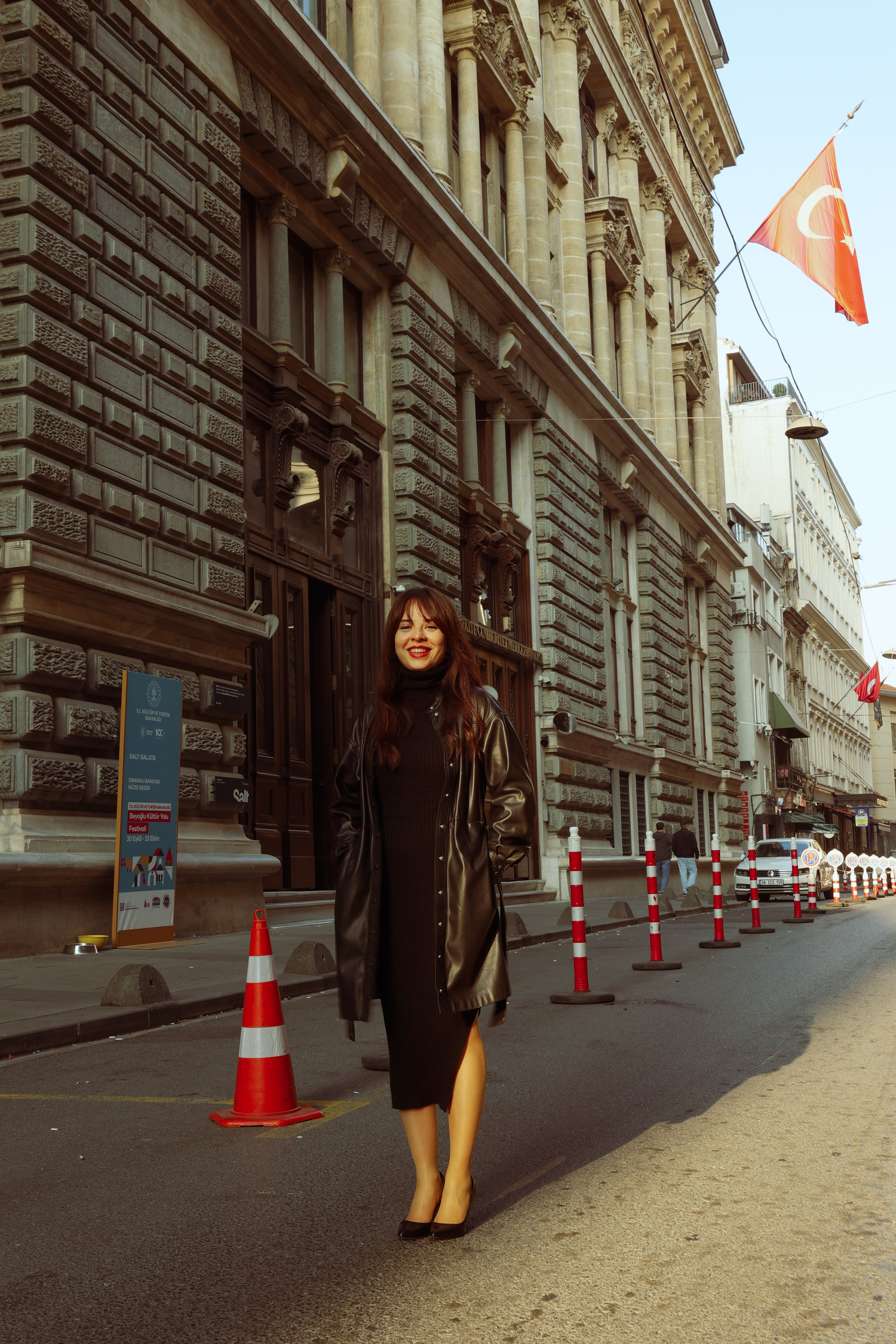 A fashionable woman in a black dress poses happily on an urban street with a Turkish flag in the background.