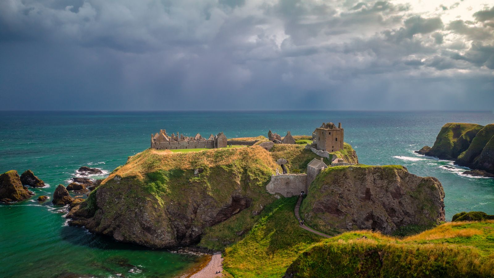 Aerial drone view of the ruins of Dunnottar Castle on a rocky sea headland under a stormy sky in Scotland, an illustrative real photo chosen to echo the cliffside strongholds of Westeros
