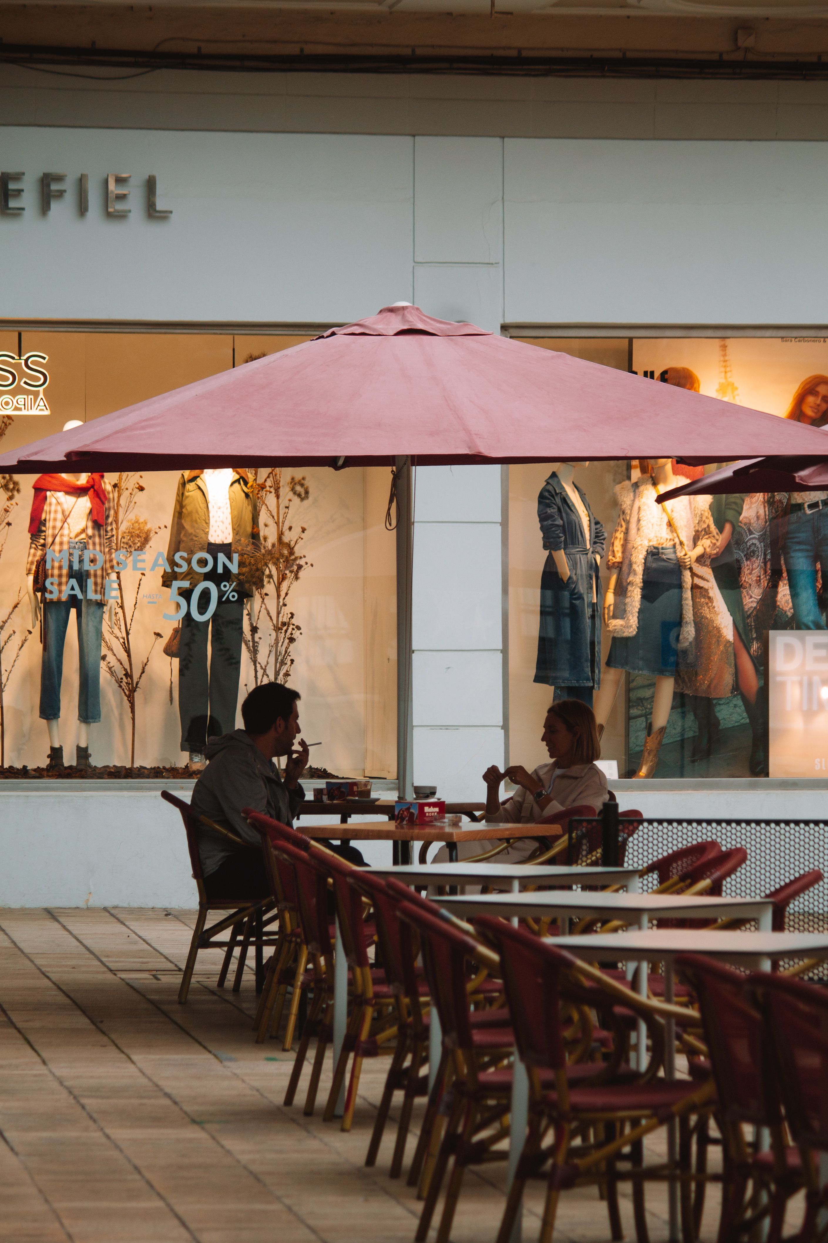 A couple conversing at a café under a large umbrella, set against a shop window backdrop.