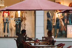 A couple conversing at a café under a large umbrella, set against a shop window backdrop.