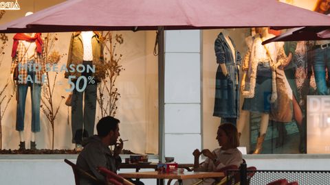 A couple conversing at a café under a large umbrella, set against a shop window backdrop.