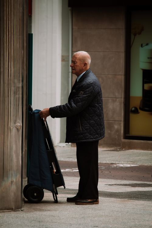 Free Senior man in jacket with cart standing on a city sidewalk. Urban street scene. Stock Photo