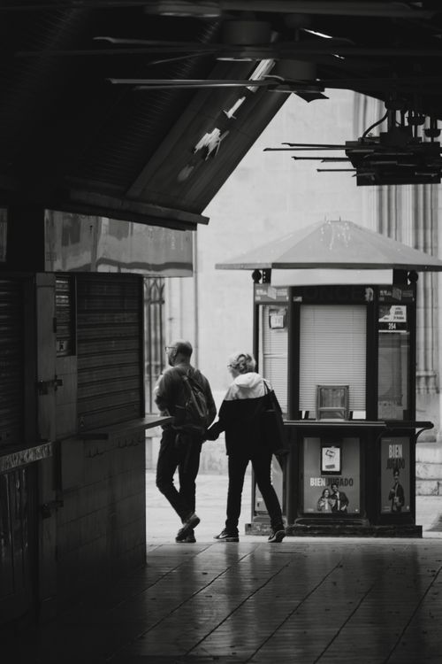 Free Black and white urban shot of two people walking by a kiosk in an alley. Stock Photo