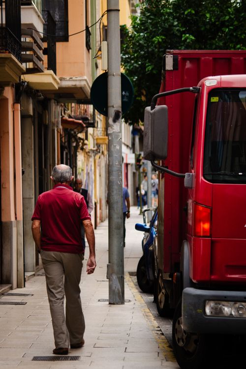 Free A senior man walking on a narrow city street alongside parked vehicles. Stock Photo