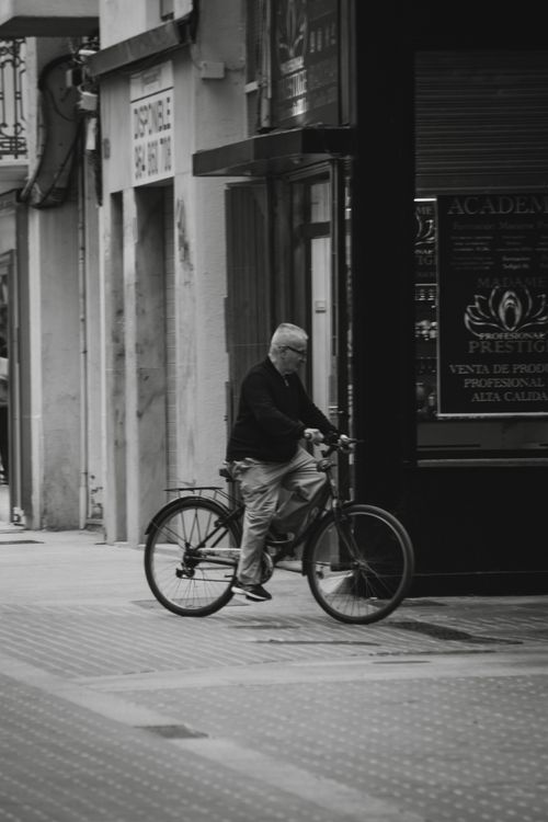Free Black and white photo of an elderly man riding a bicycle in a city street, showcasing urban life. Stock Photo