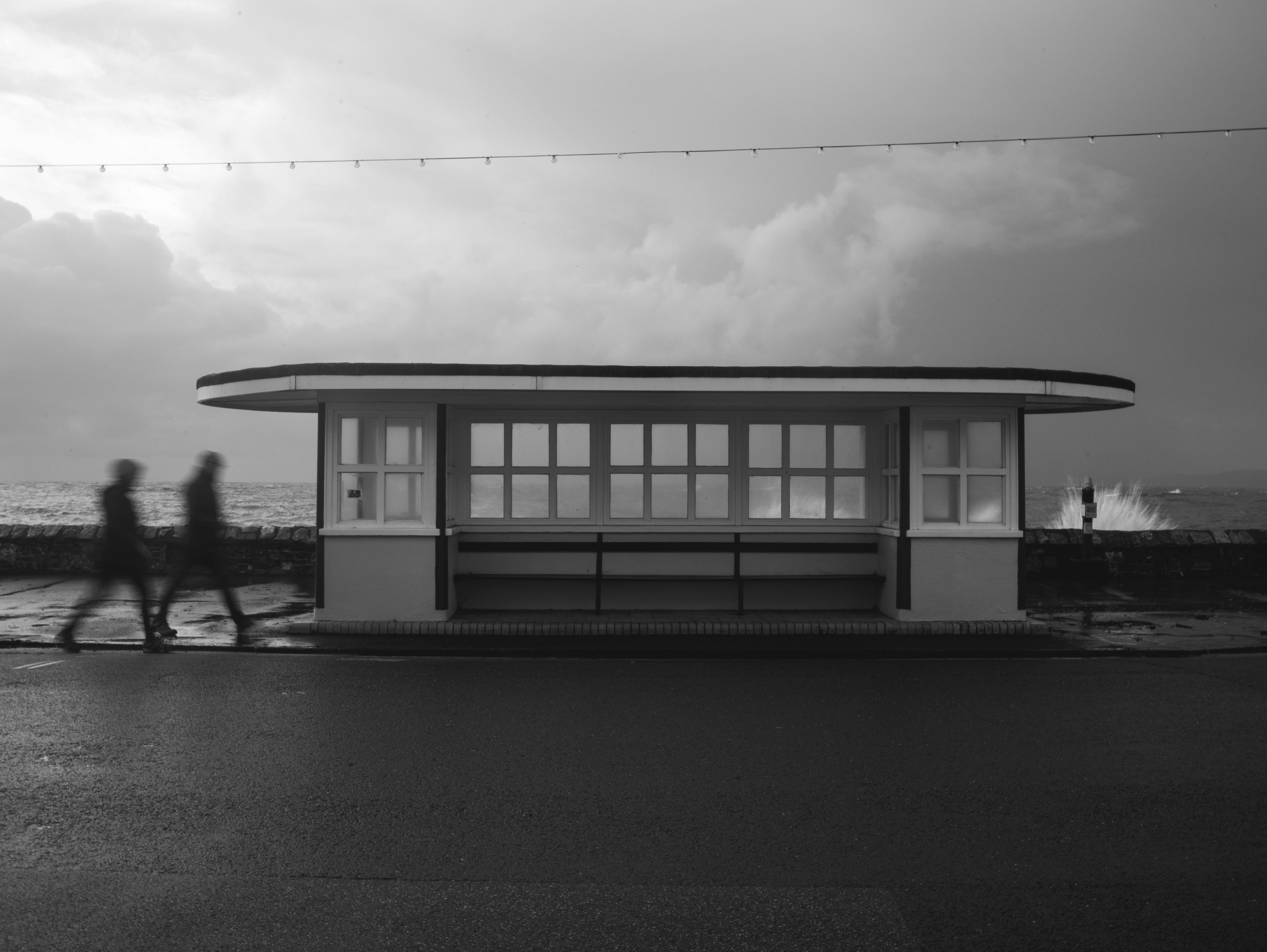 Monochrome image of a modernist waterfront shelter with blurred figures walking by.