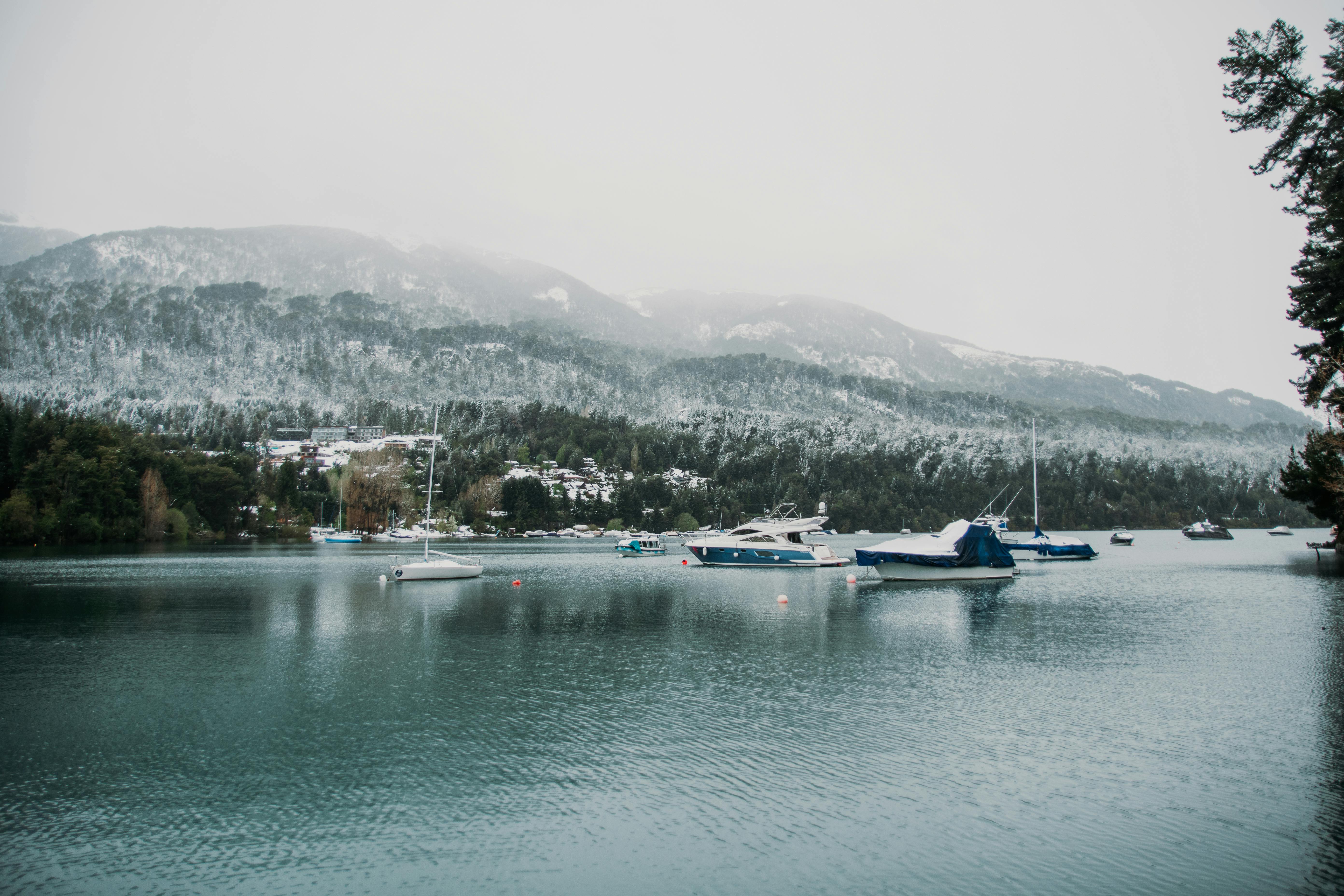 Tranquil winter scene of boats on a lake surrounded by snowcapped mountains and forested shoreline.
