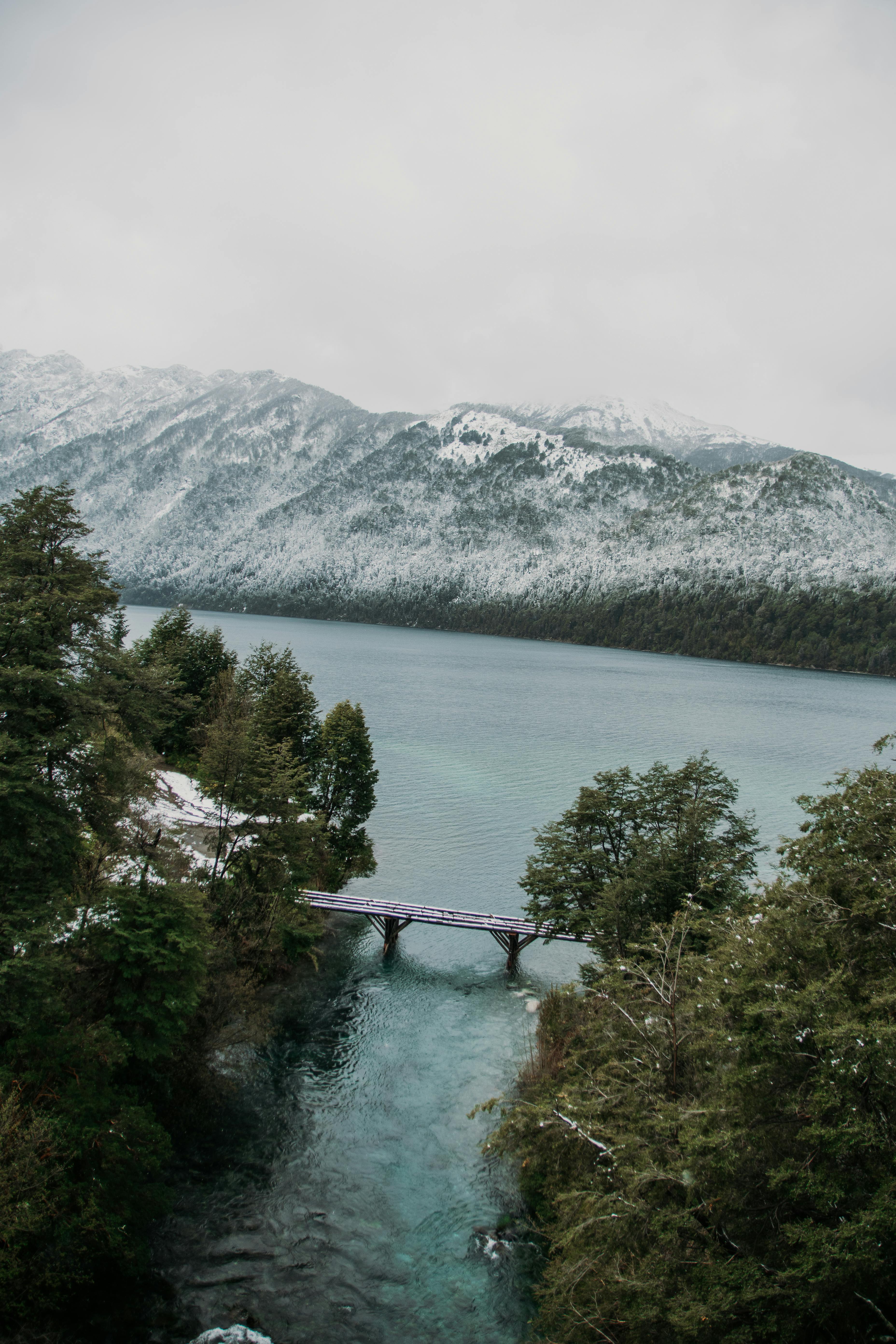 Panoramic view of Correntoso Lake with a bridge, surrounded by snowy mountains and lush trees.