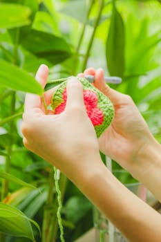 Close-up of hands crocheting a red and green piece surrounded by vibrant green foliage.