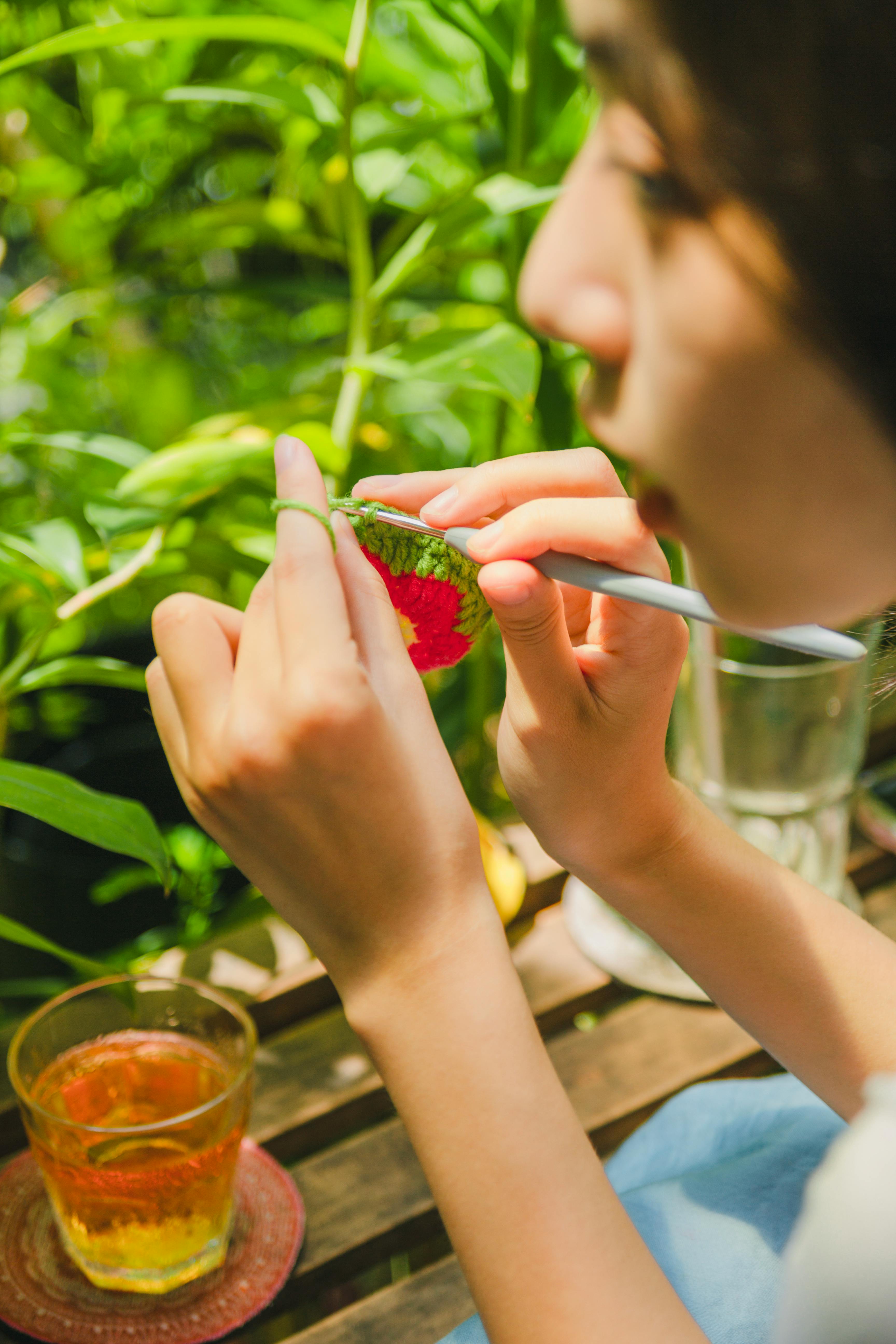 Close-up of a woman crocheting in a serene garden setting with herbal tea on a sunny day.