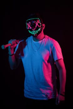 Man wearing a neon LED Halloween mask holding a baseball bat in a dark studio setting.