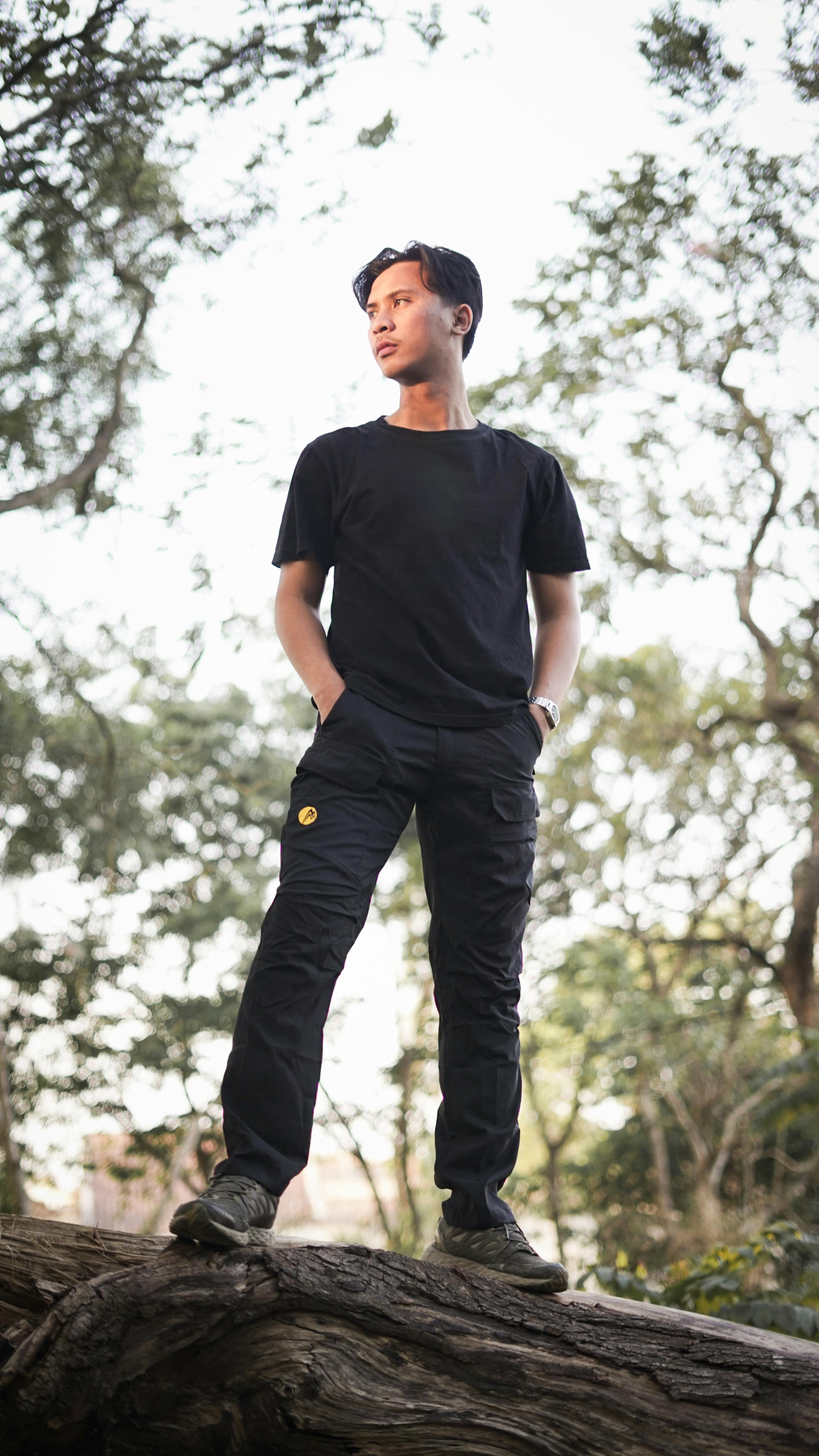 A young man stands confidently on a log in a scenic park, surrounded by trees.