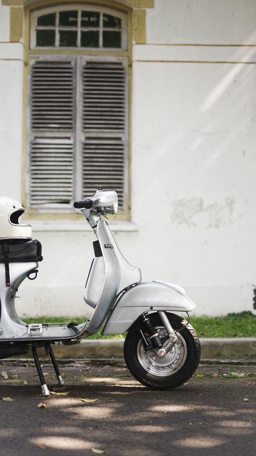 Free A vintage white scooter parked next to a rustic window with shutters on a quiet city street. Stock Photo