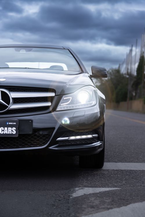 Free Close-up of a Mercedes car with headlights on, driving on an urban road under cloudy skies. Stock Photo
