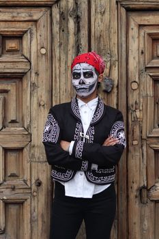 A man wearing skull face paint and traditional attire stands against a wooden door for Dia de los Muertos.