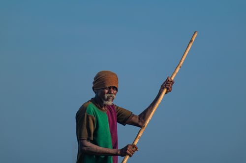 Free An elderly man with a headscarf holds a wooden paddle against a clear blue sky, creating a striking outdoor portrait. Stock Photo