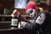 Person with skeleton face paint in traditional Mexican attire holding Creyente bottle at a bar.