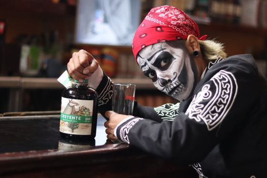 Person with skeleton face paint in traditional Mexican attire holding Creyente bottle at a bar.