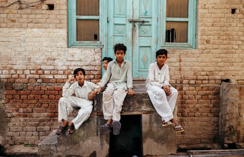 Free Four boys in traditional outfits sit by a rustic brick wall and turquoise door, enjoying a leisurely day outdoors. Stock Photo