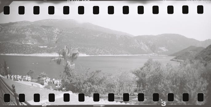Vintage black and white photo of a serene seascape view captured through a filmstrip perspective, with mountains and trees lining the shore.