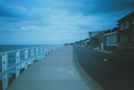 A serene coastal urban street at twilight, with ocean views and few people walking.