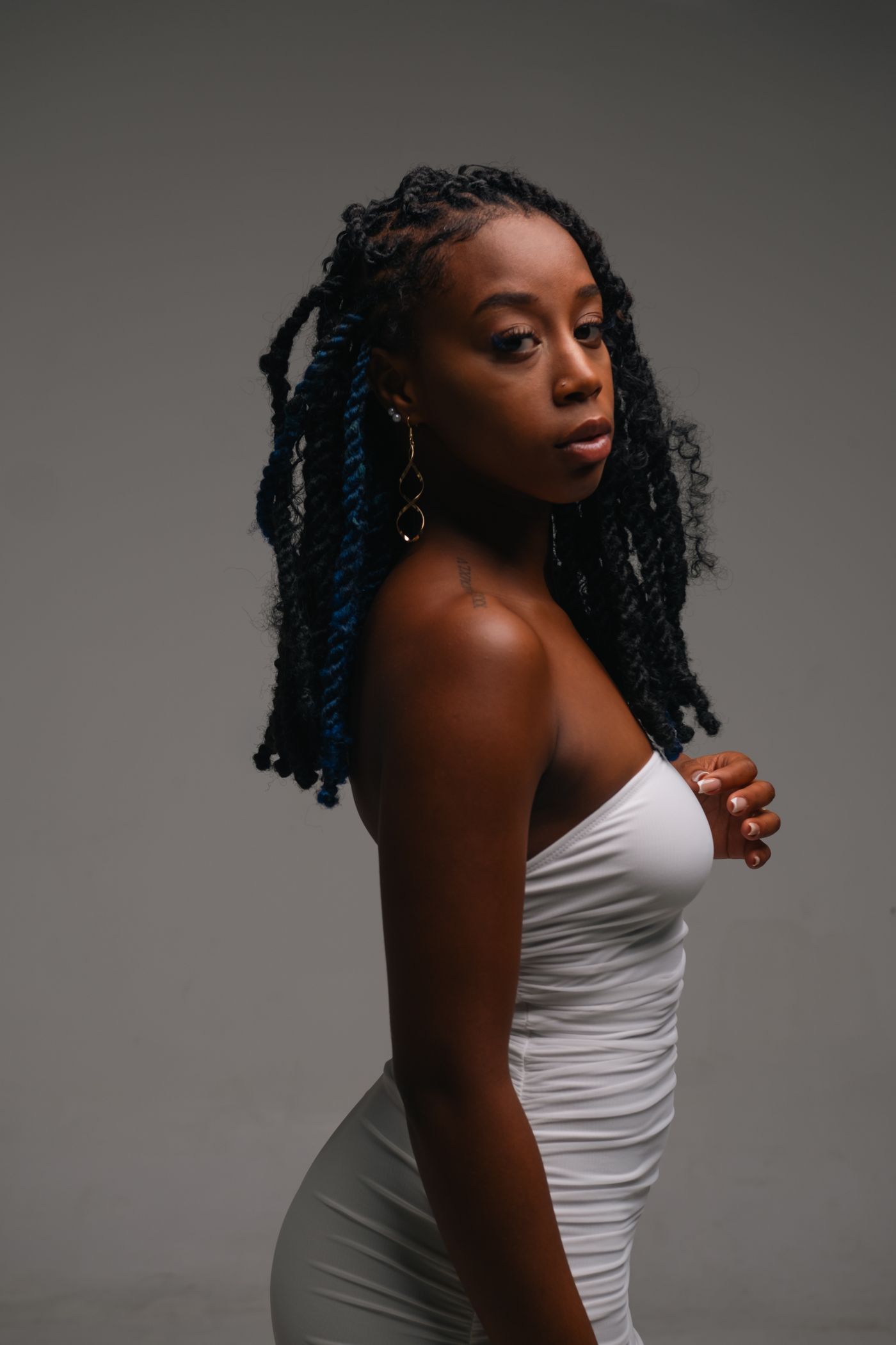 A woman in a white ruched dress, gold earrings, photographed in studio light.