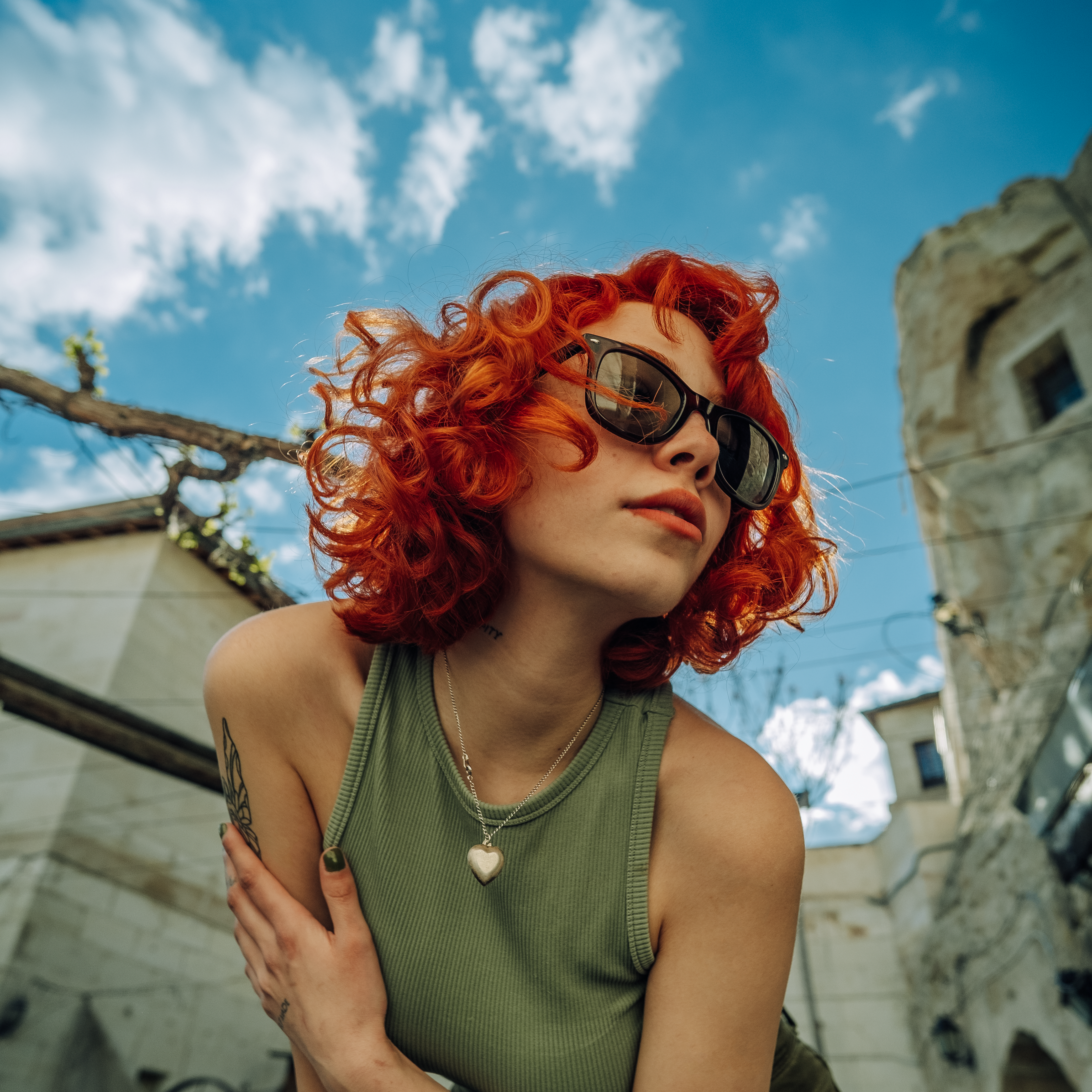 Young woman with curly red hair and sunglasses posing outdoors in a green tank top.
