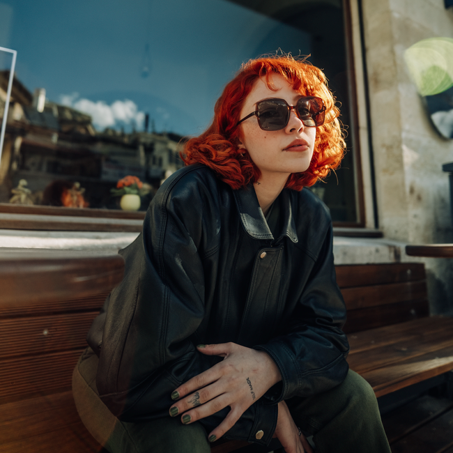 Redhead woman in leather jacket posing confidently outside a window, exuding modern fashion vibes.