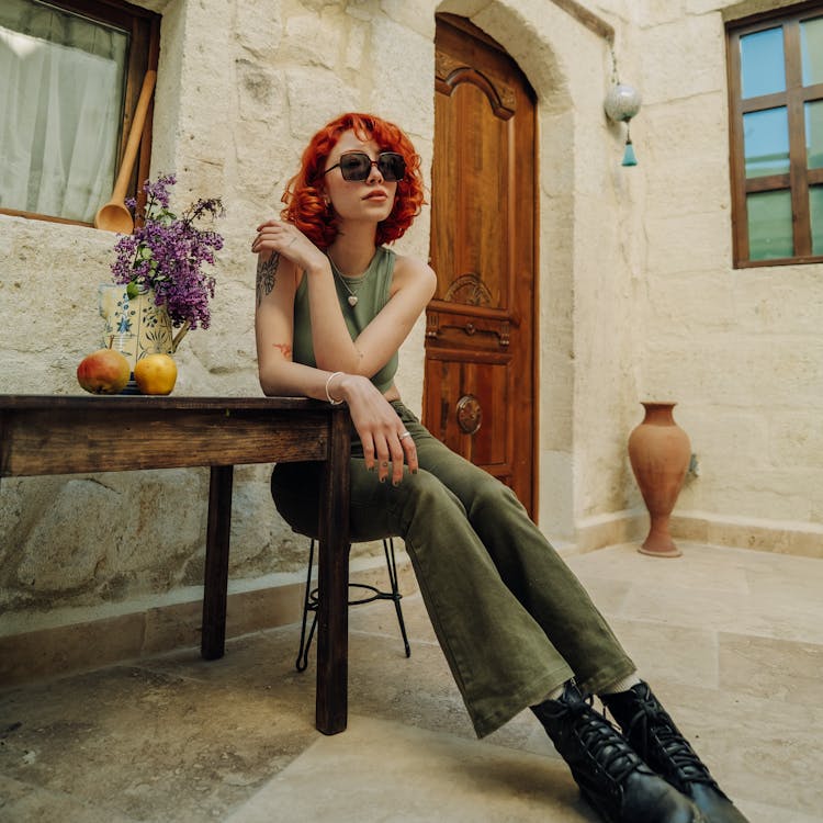 Young Redhead In Sunglasses Sitting At The Table Outside Of A House 