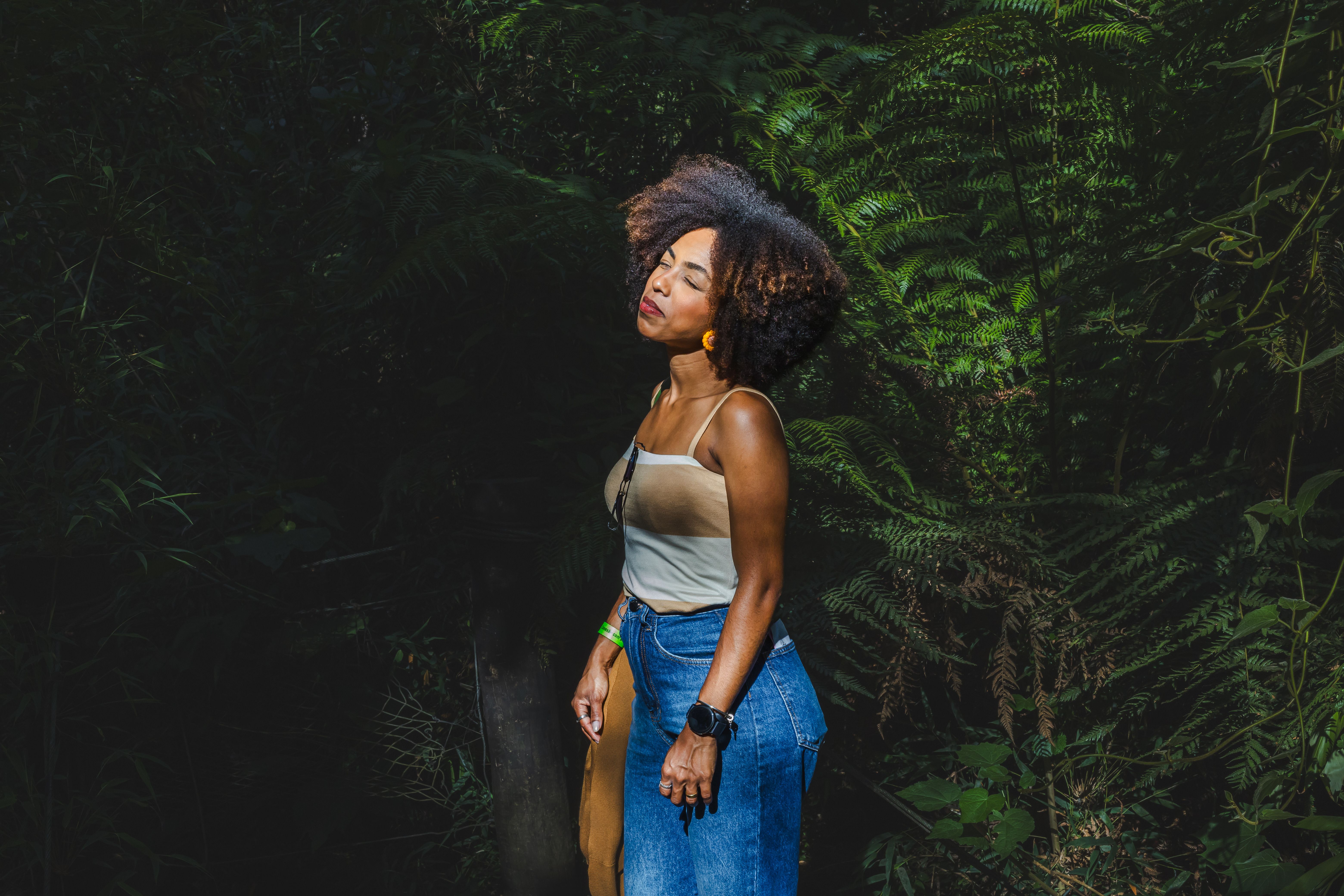 A fashionable woman enjoys the summer sun surrounded by lush ferns, wearing casual attire in an outdoor setting.