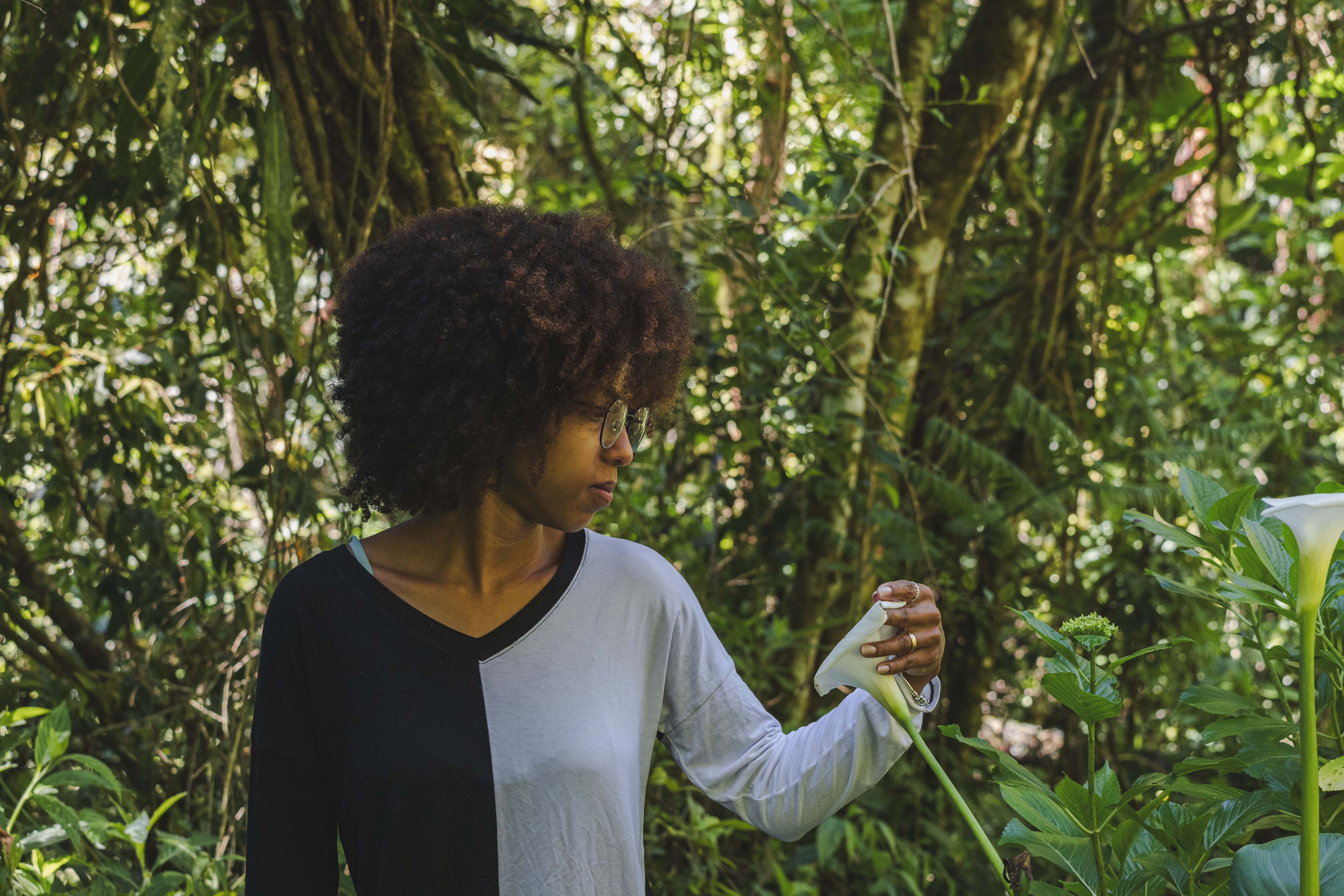 A woman with curly hair and eyeglasses stands amidst a lush forest holding a white flower.