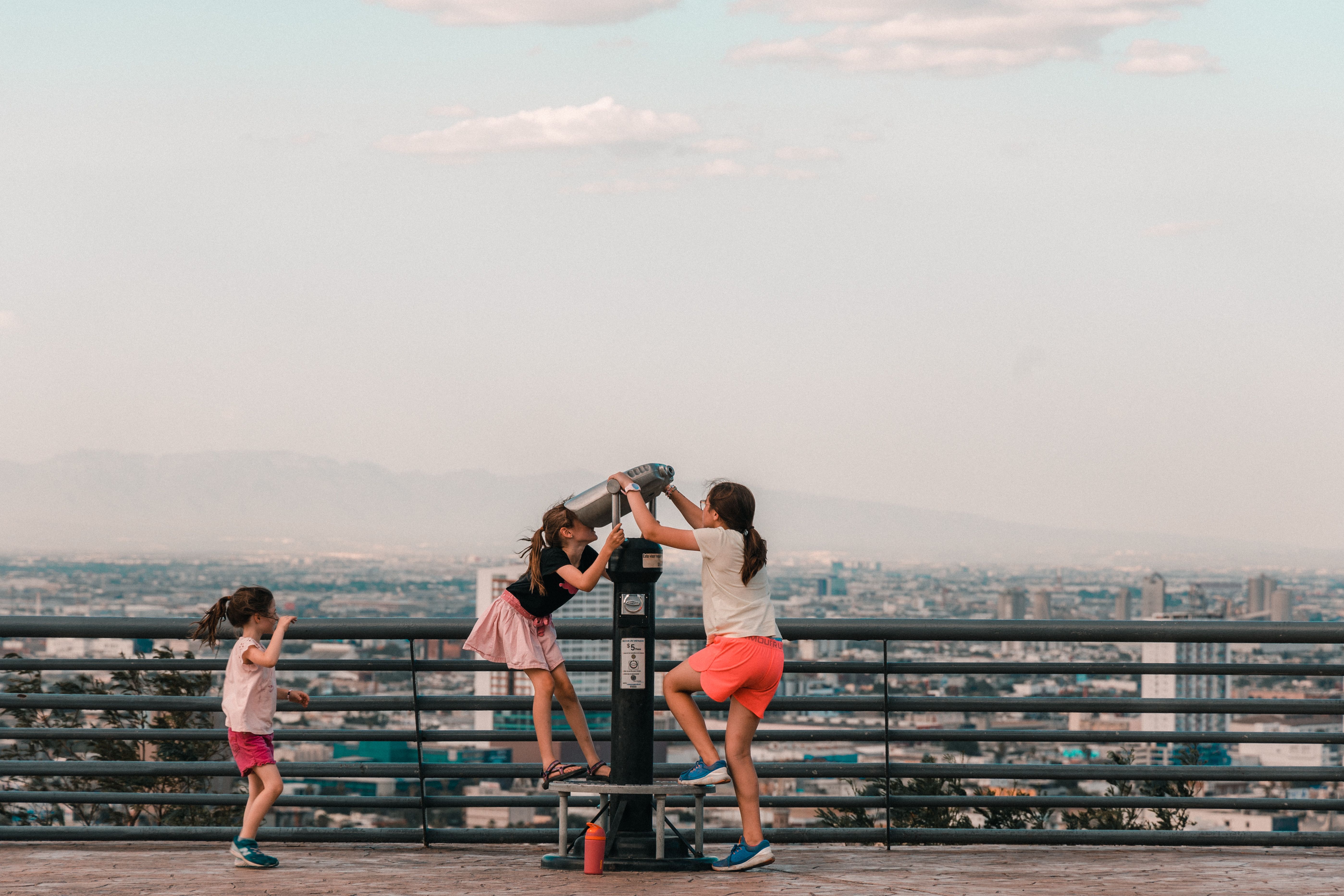 Three girls enjoy viewing a cityscape through a telescope from a scenic overlook, capturing childhood wonder.