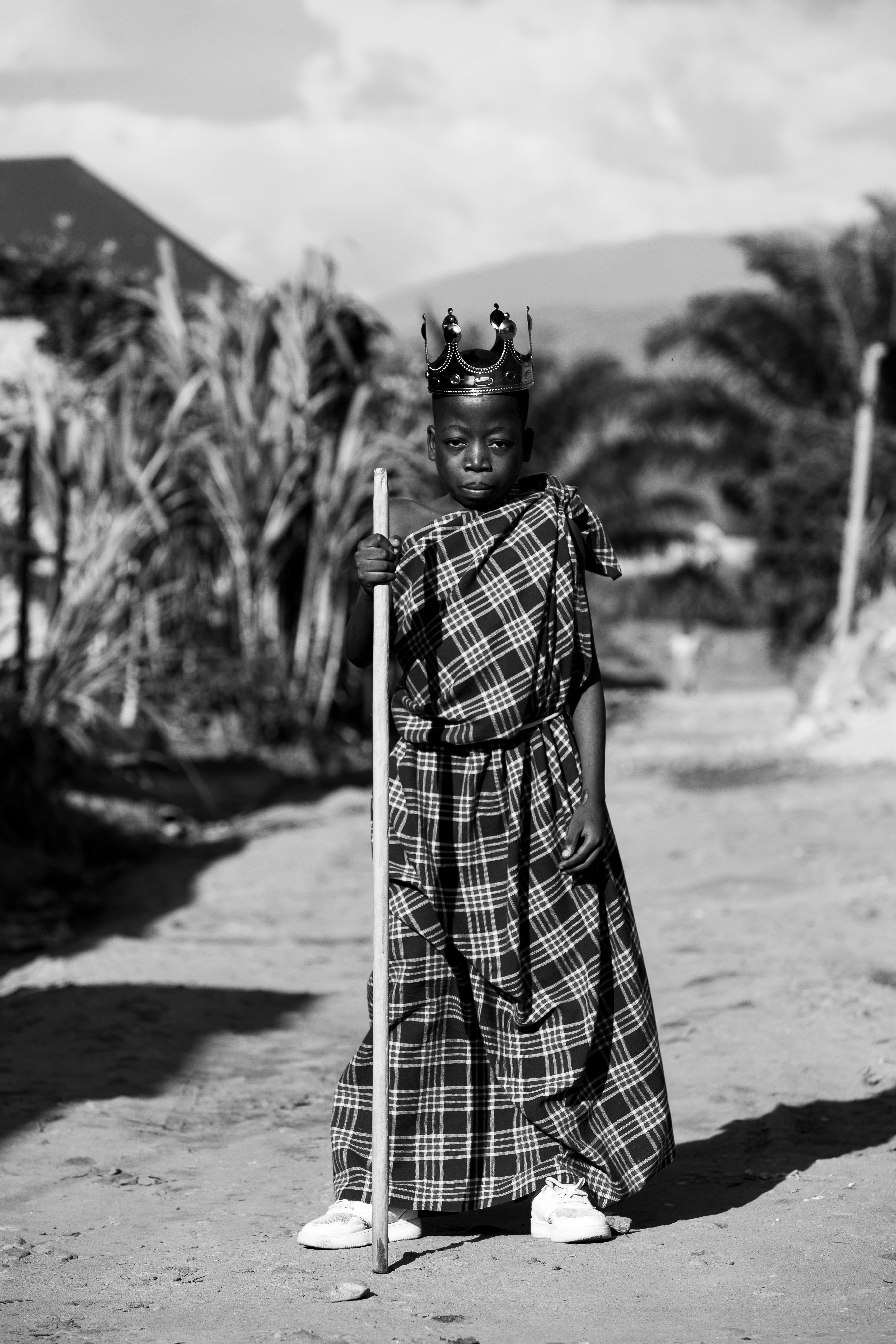 Black and white portrait of a child in traditional attire with a crown, holding a stick outdoors.