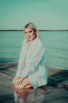 Blonde woman kneeling on beach with turquoise sea in background, wearing a white shirt.