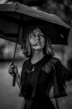 Black and white portrait of a woman holding an umbrella on a rainy city street.