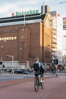Cyclist rides past Heineken Brewery in bustling Amsterdam city street.